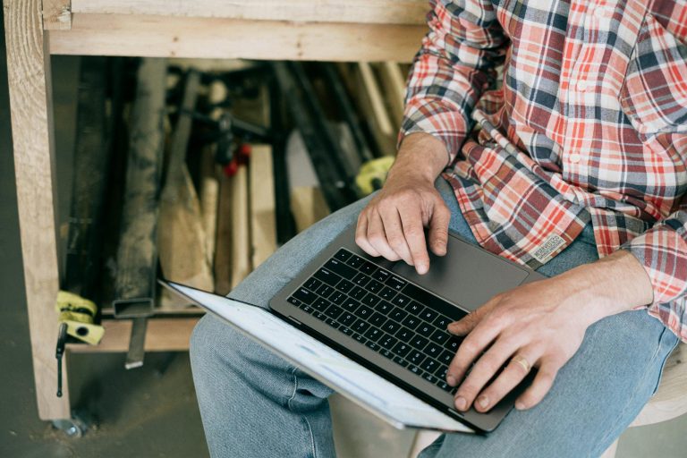 Craftsman working on laptop in woodworking workshop, surrounded by tools.